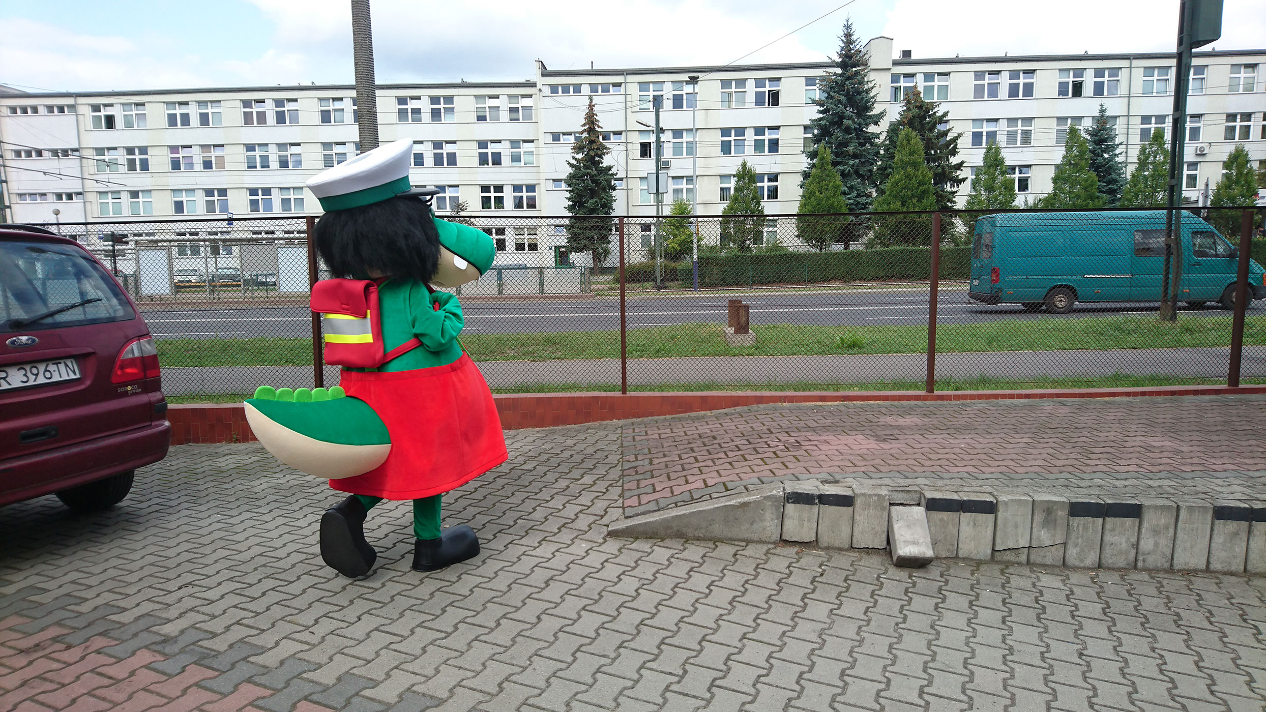 Na fotografii widnieje kostium krokodyla z długimi czarnymi włosami czapką ITD, w sukience i z tornistrem na plecach, przemierzający krakowskie ulic. Po jezdni poruszają się samochody, w tle budynek biurowy MPK w Krakowie